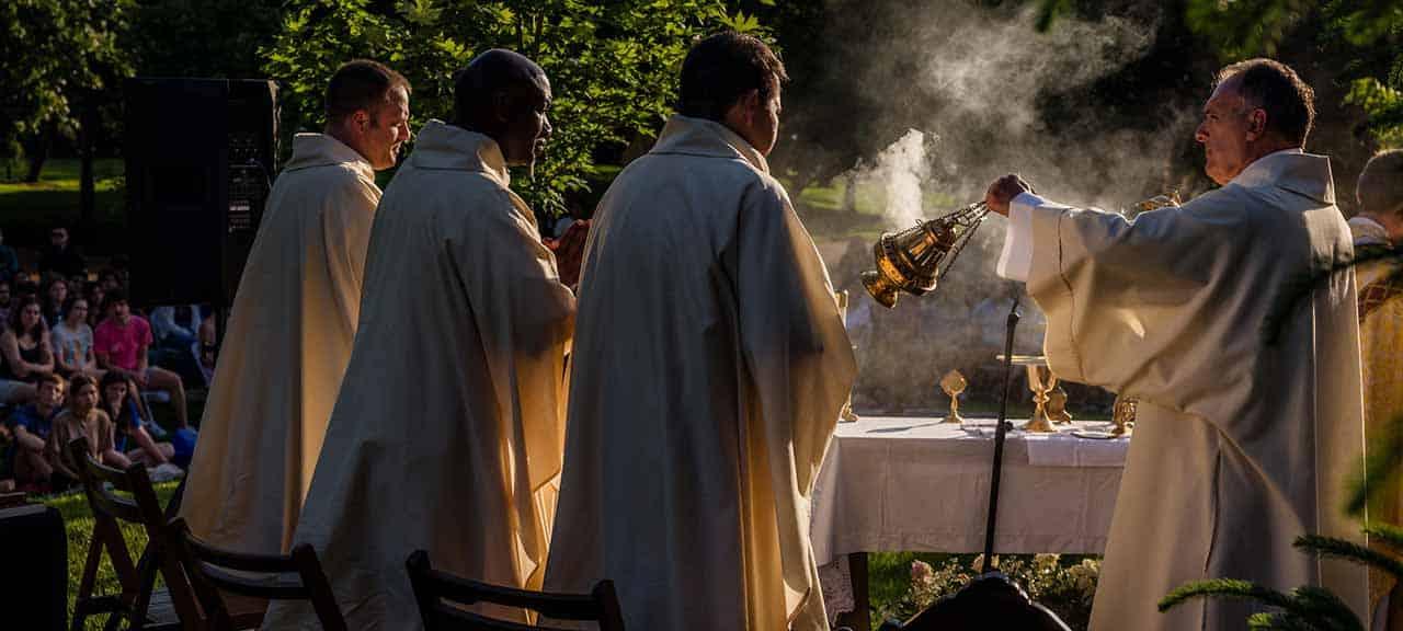 A man wearing white vestments swings a censer over the alter while celebrating Mass. Three other men in vestments look on.