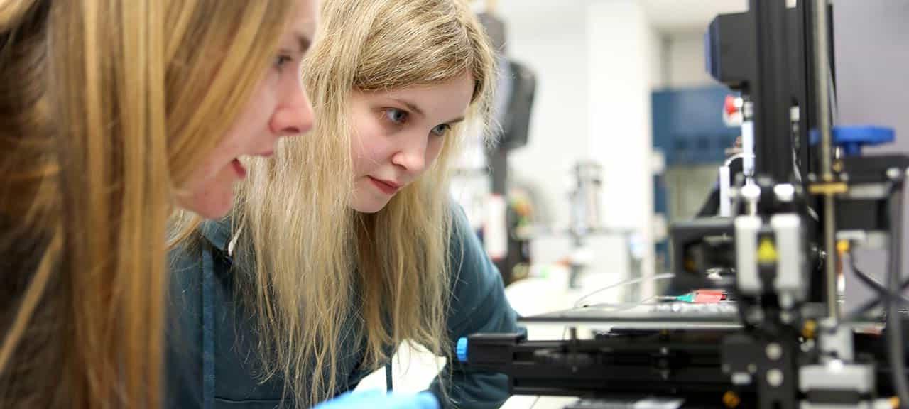 Two female students with long blonde hair lean in close to study a piece of equipment in a laboratory.